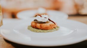 A close-up view of a gourmet dish featuring a golden patty topped with cream and a chili, served on a green sauce