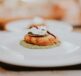 A close-up view of a gourmet dish featuring a golden patty topped with cream and a chili, served on a green sauce